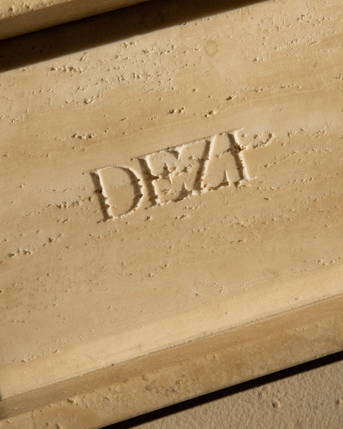 This shows a closeup of travertine stone tray with the DEZI logo carved into it. Sunlight is shining on the tray, casting beautiful shadows.