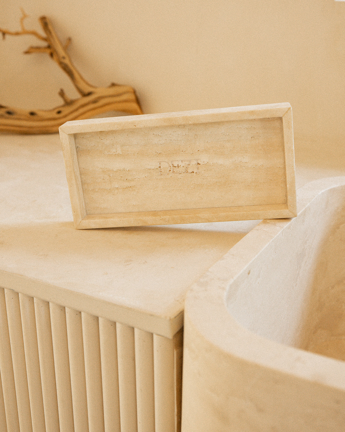 This shows a travertine stone tray sitting on a counter with the DEZI logo carved into it. Sunlight is shining on the tray, casting beautiful shadows.