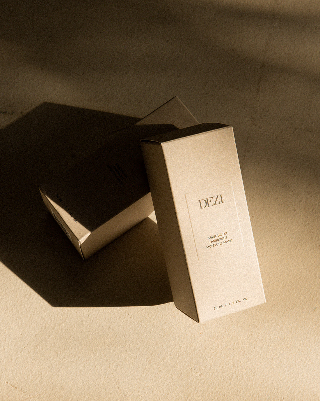 This shows two DEZI SKIN moisture mask boxes leaning on each other. The sunlight is hitting them, casting long dark shadows on the stone floor. 