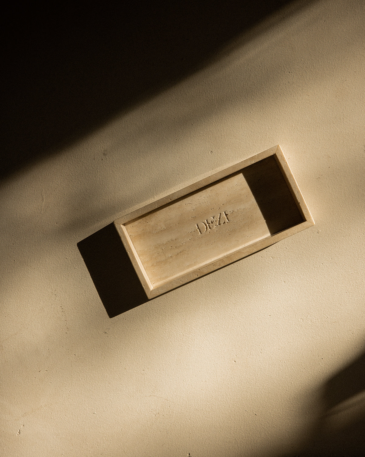 This shows a travertine stone tray with the DEZI logo carved into it. Sunlight is shining on the tray, casting beautiful shadows. 