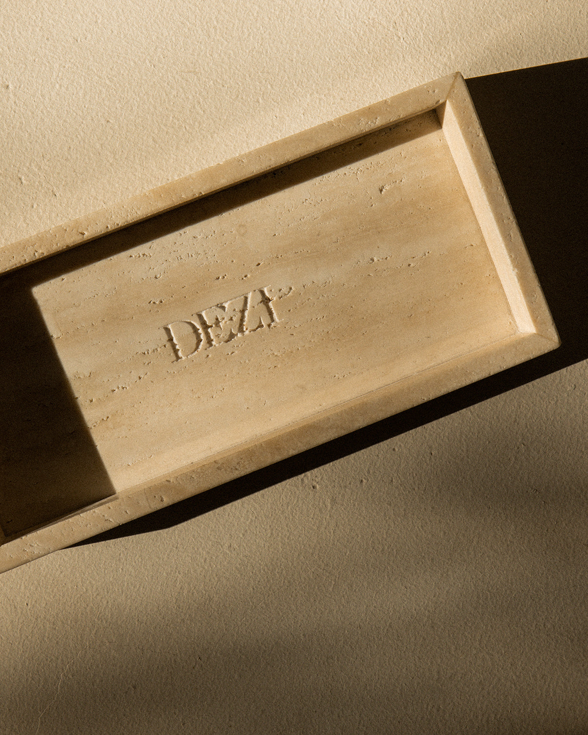 This shows a travertine stone tray with the DEZI logo carved into it. Sunlight is shining on the tray, casting beautiful shadows. 