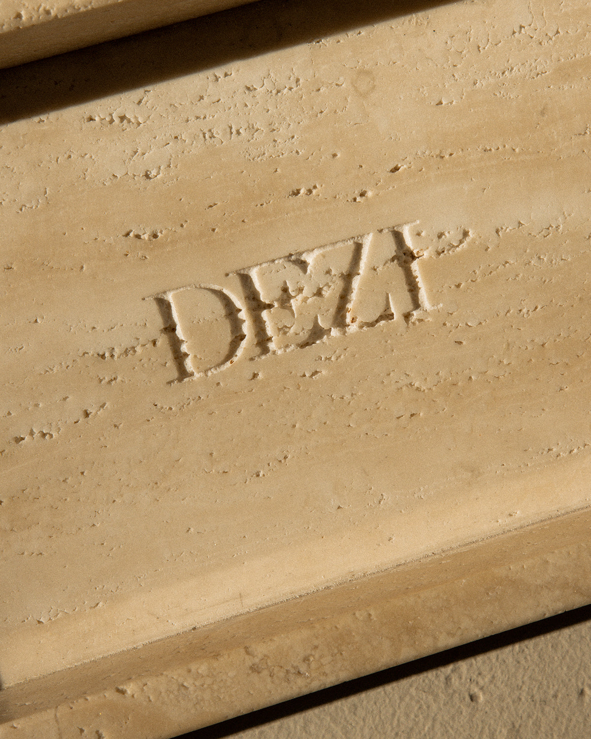 This shows a closeup of travertine stone tray with the DEZI logo carved into it. Sunlight is shining on the tray, casting beautiful shadows. 