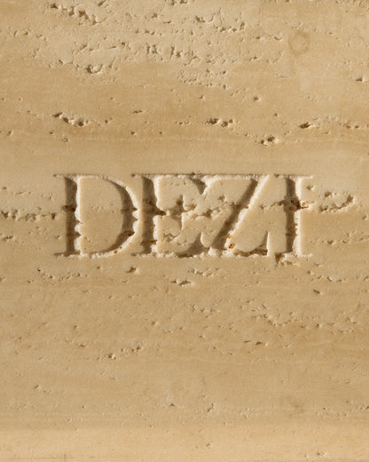 This shows a closeup of travertine stone tray with the DEZI logo carved into it. Sunlight is shining on the tray, casting beautiful shadows. 