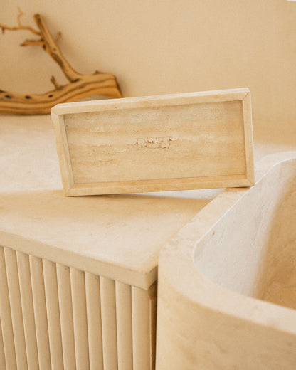 This shows a travertine stone tray sitting on a counter with the DEZI logo carved into it. Sunlight is shining on the tray, casting beautiful shadows. 